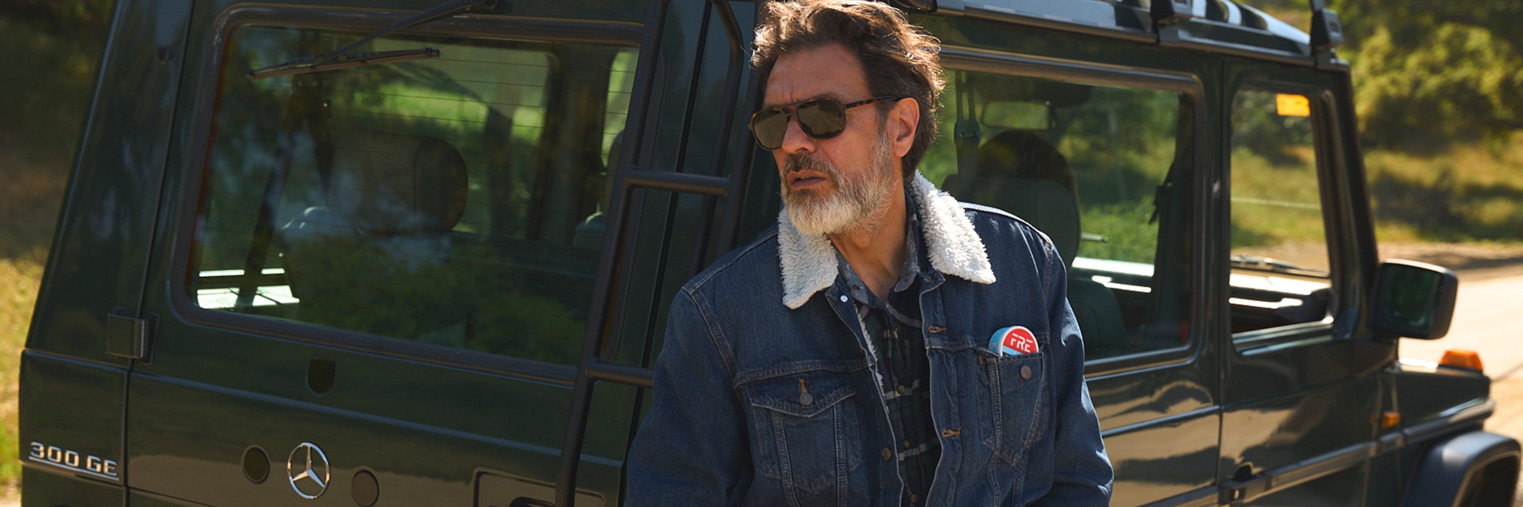 Bearded man in sunglasses with FRE Pouch tin in jacket pocket, posed beside a Mercedes 300GE, highlighting bold and adventurous lifestyle