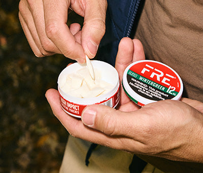 Hands opening a FRE Wintergreen 12 mg mega pouch tin, showing white nicotine pouches inside