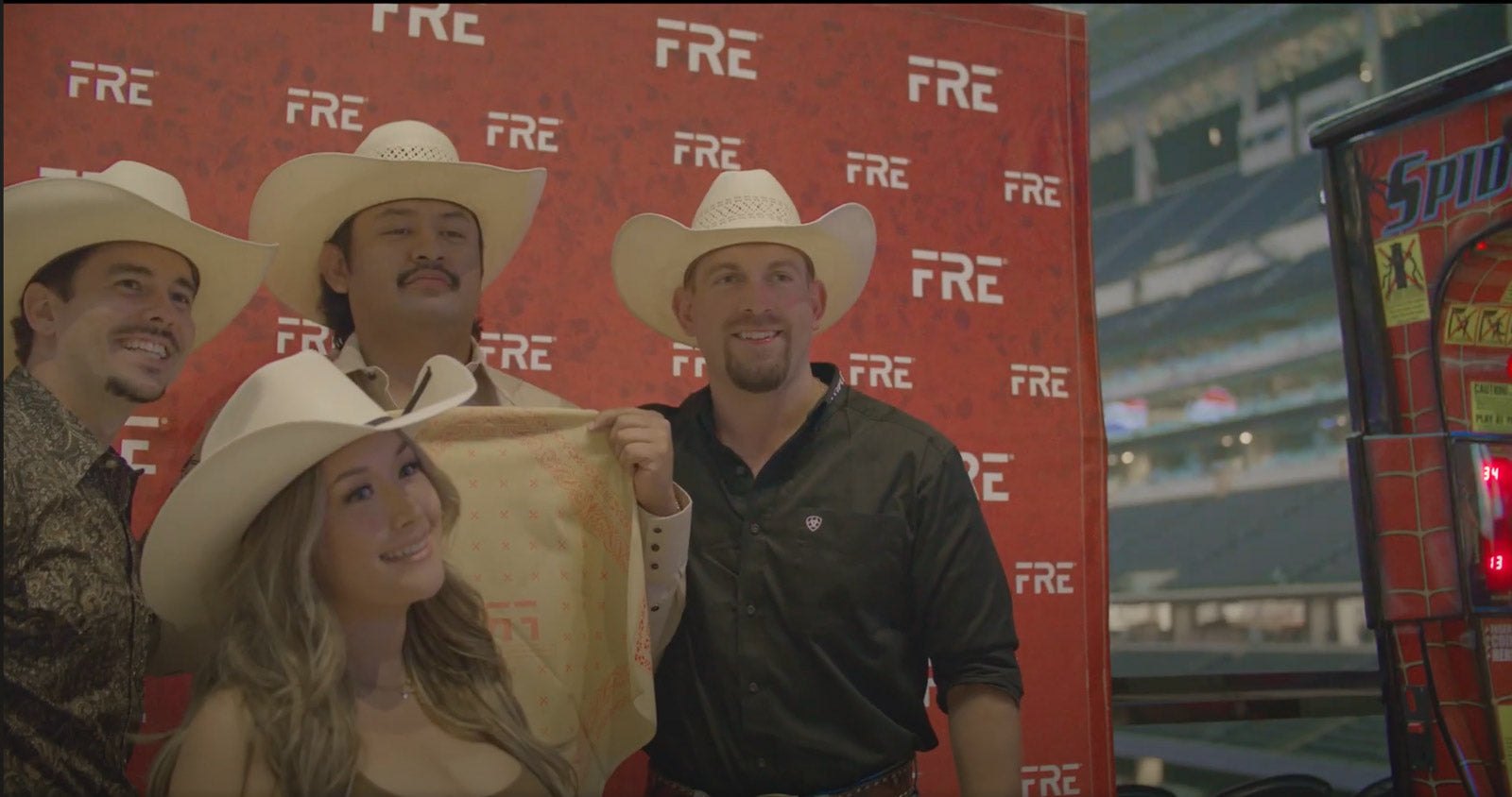 Group of smiling people in cowboy hats posing in front of a FRE step-and-repeat banner at an event, with one holding a branded bandana and a Spider-Man arcade game in view.