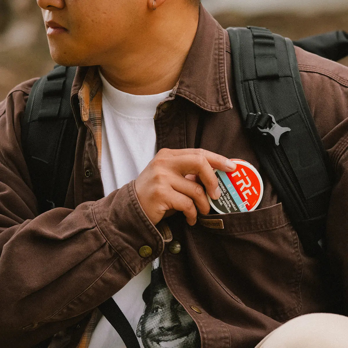 Person sliding a round nicotine pouch tin labeled FRE into a jacket chest pocket, with a backpack strap across the shoulder, emphasizing portability and everyday use